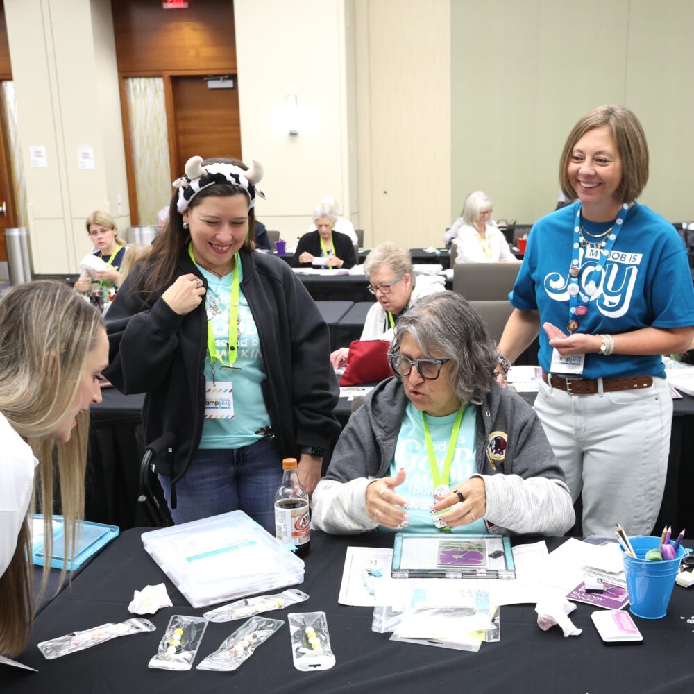attendees at a craft workshop table with beads, paper, and tools; a woman wearing a cow-themed headband smiles on the left, another woman in a blue shirt stands nearby with a badge.
