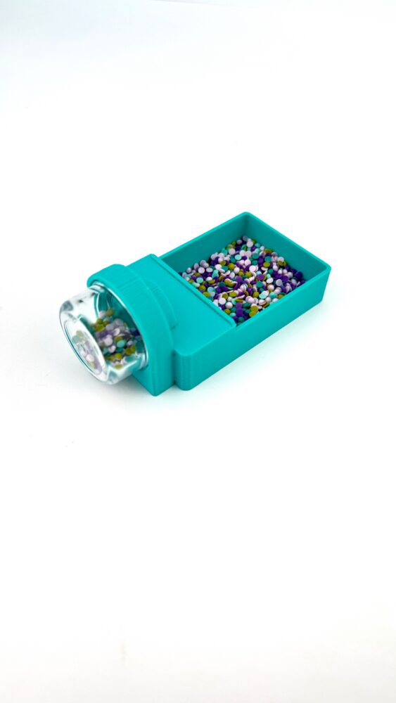 Teal plastic jar with a clear cap tipped into a matching teal container filled with multicolored round candy sprinkles (purple, white, green, yellow).