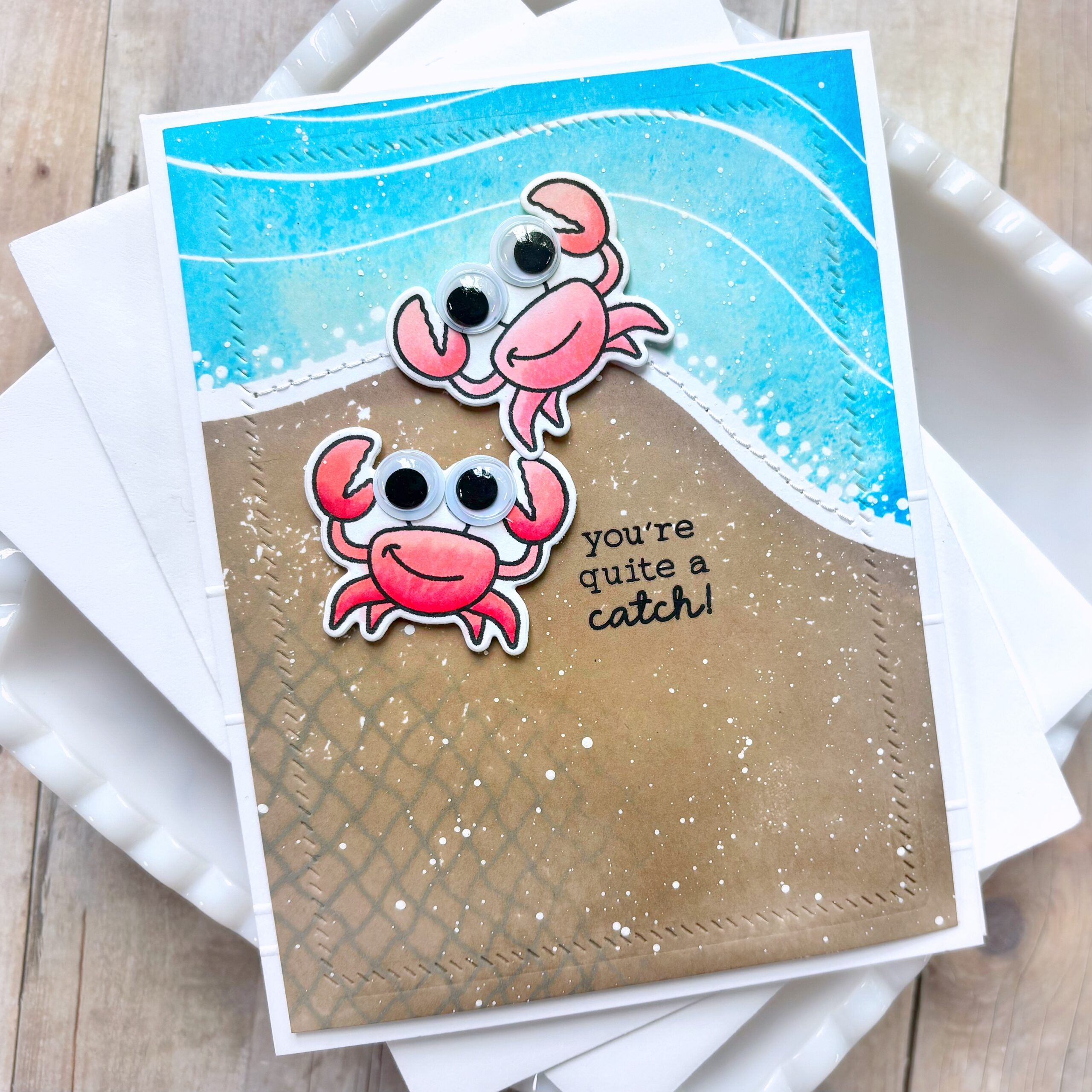 Stack of handmade greeting cards featuring pink crabs with googly eyes on blue ocean-themed backgrounds on a wooden surface.