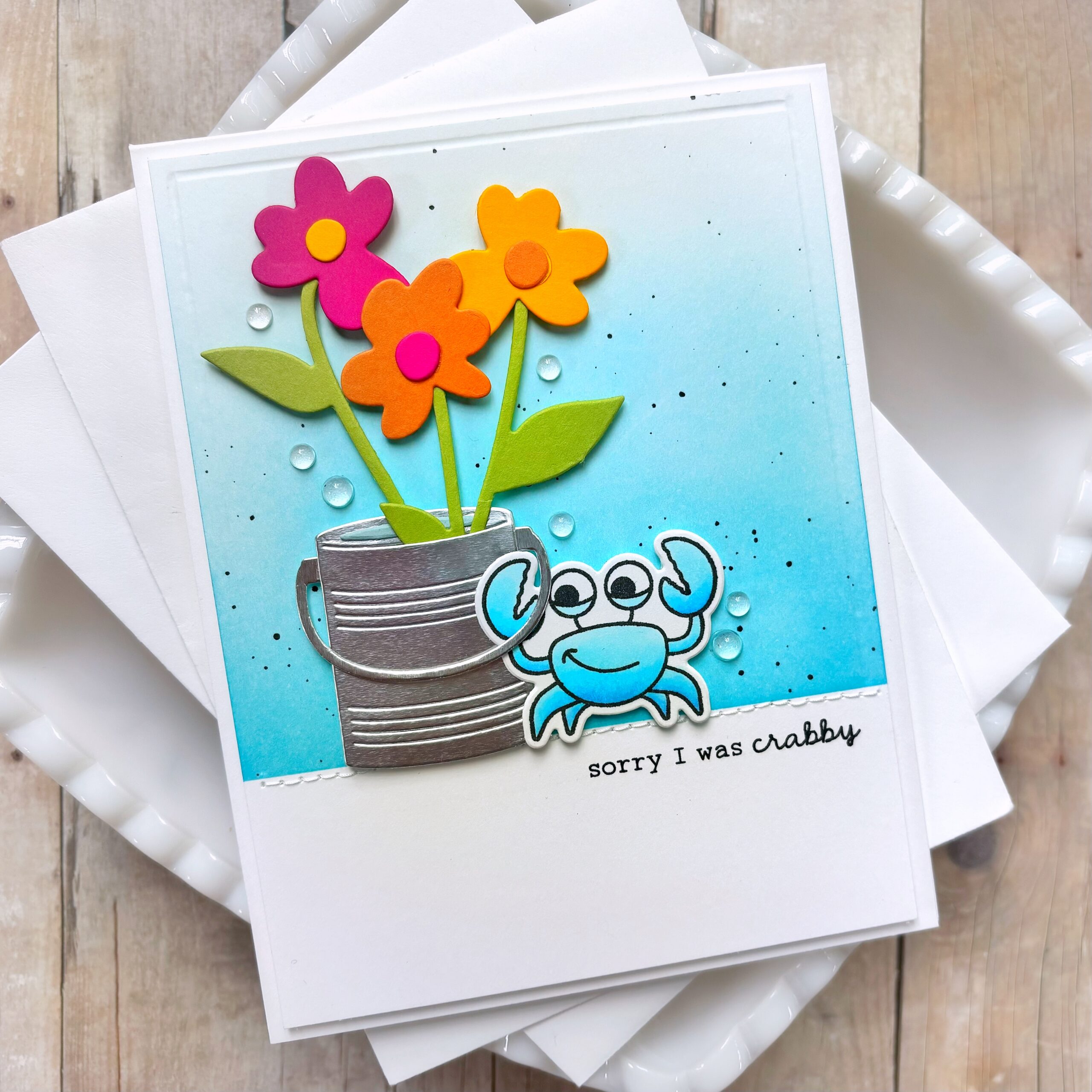 Bright handmade greeting card featuring pink and orange paper flowers in a pot on a blue gradient background, with white envelopes underneath and decorative water droplets motif while stacked.