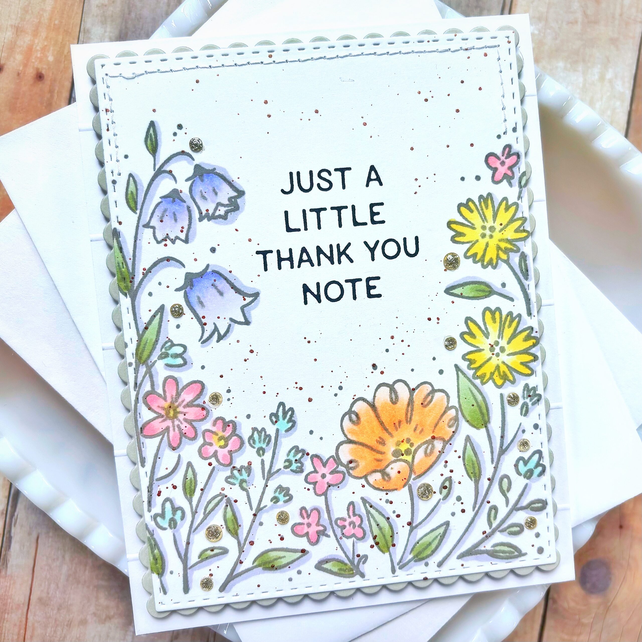 Handmade thank-you card with colorful flowers, scalloped edge, and gold glitter on a wooden surface.