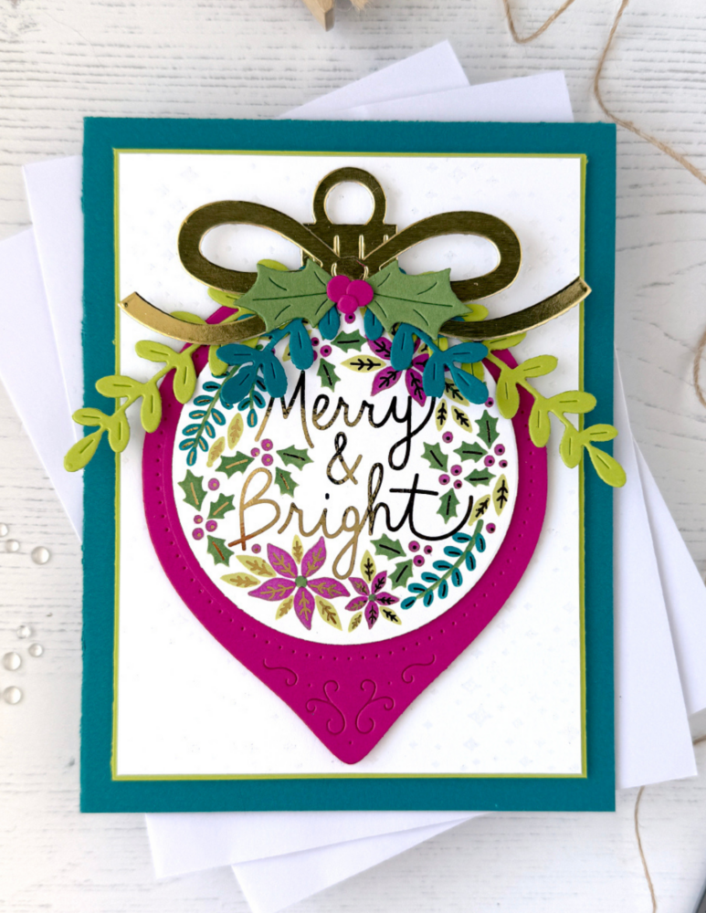 Handmade Christmas card with gold bow accent, green holly leaves, and a colorful floral wreath surrounding the 'Merry & Bright' greeting